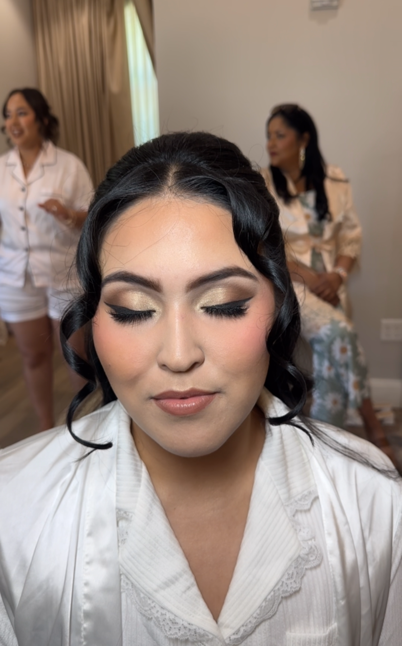 Bride in a white robe with makeup application in a room with other people.